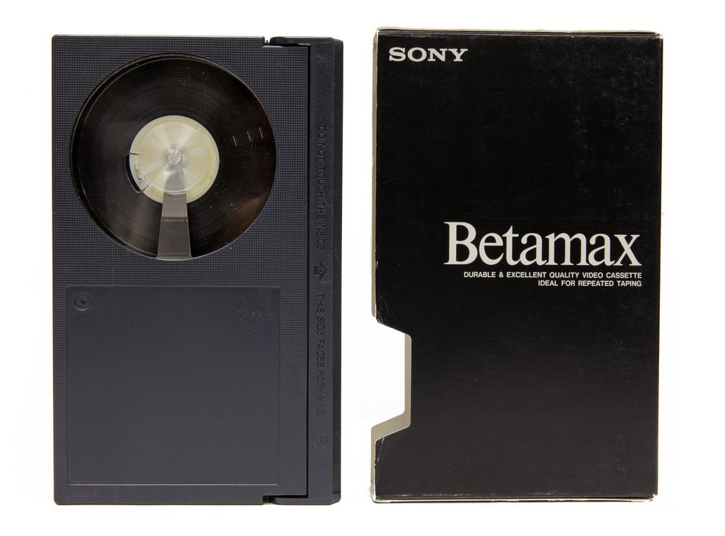 A Betamax videocassette positioned on the left, with the cardboard case reading "Betamax, durable &excellent quality video cassette, ideal for repeated taping" positioned on the right.